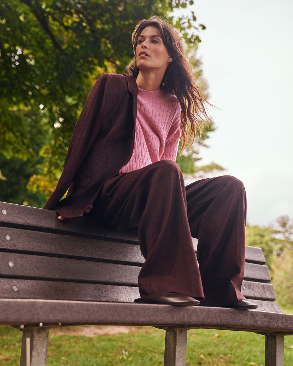 A woman sitting on a park bench wearing a pink cable-knit sweater, a dark brown blazer, and matching dark brown wide-leg trousers, with green trees in the background.