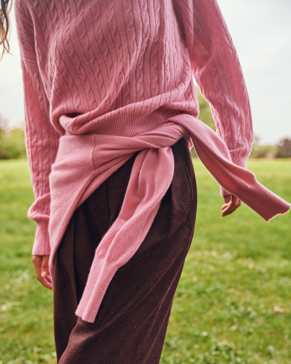 A person outdoors wearing a dusty pink cable-knit sweater tied around the waist over dark brown trousers, standing on a grassy field.