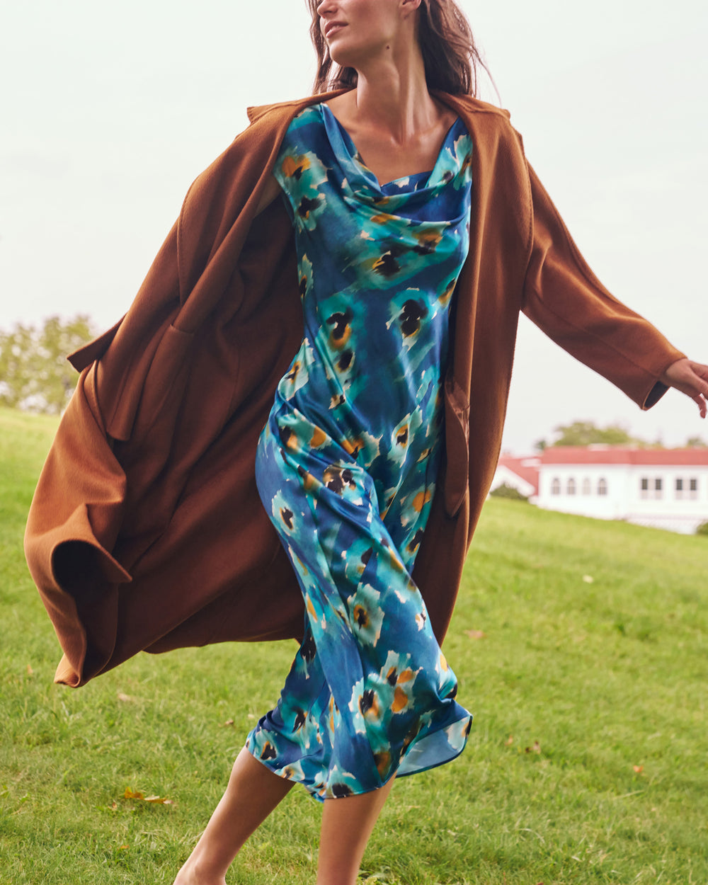 A woman outdoors on a grassy hill wearing a flowing blue floral midi dress under a draped brown coat. The background shows a soft-focus grassy field and a building in the distance.