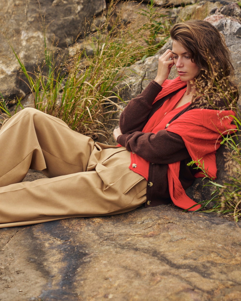 A woman reclines on a rocky outdoor surface wearing a rust-red cardigan over a brown sweater, paired with beige trousers and a red scarf.