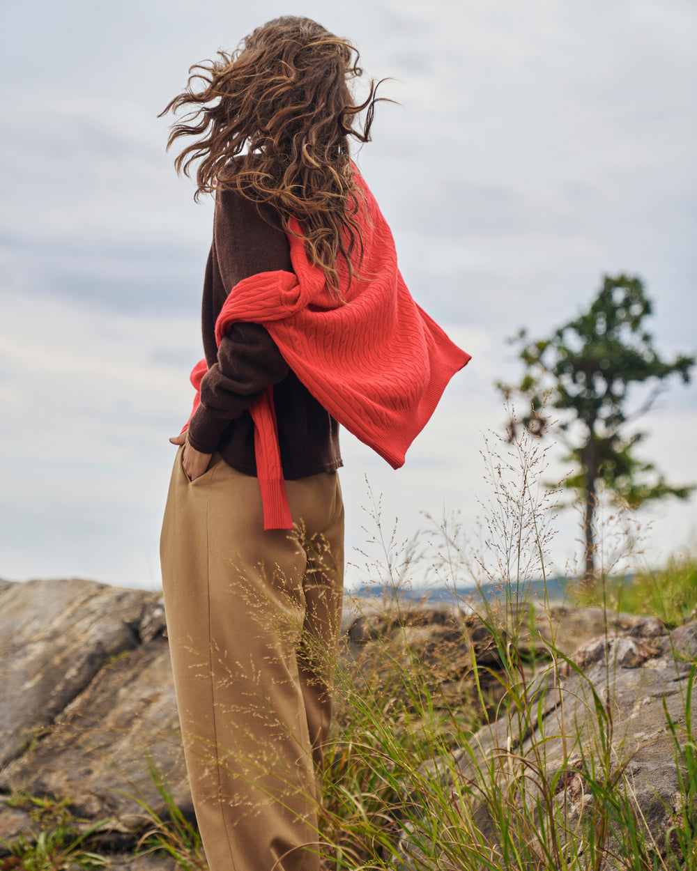 A person standing outdoors on a rocky grassy area, wearing a dark brown knit top, beige wide-leg pants, and carrying a bright coral-red knit wrap or shawl over one shoulder.