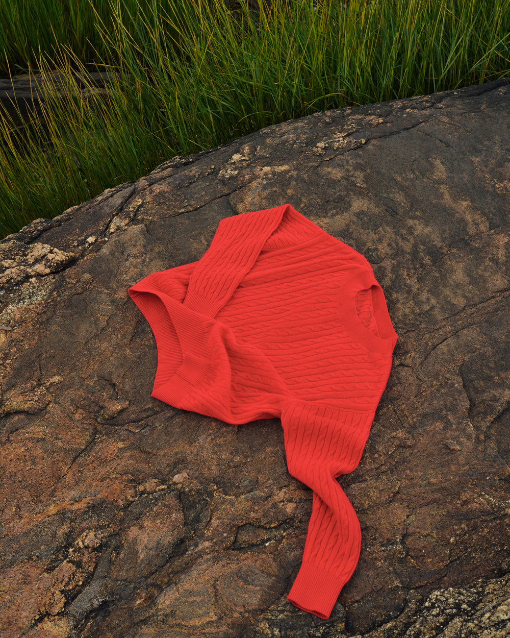 A bright coral-red ribbed knit long-sleeve top laid flat on a large textured rock with green grass in the background.