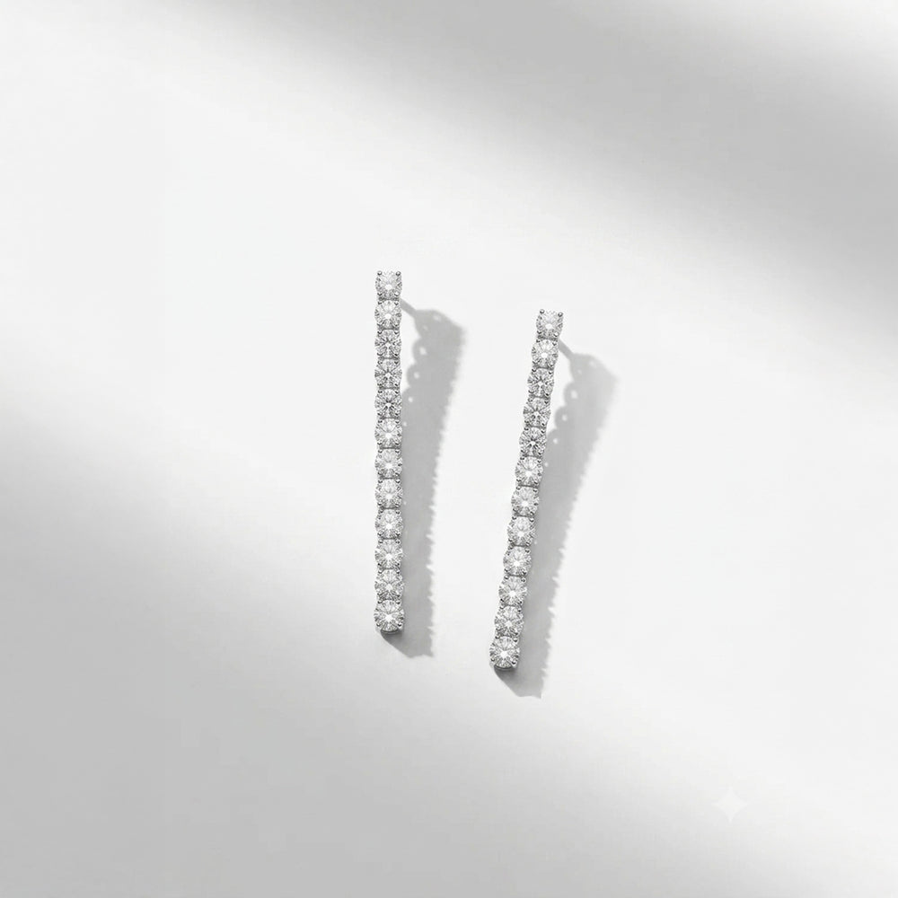 A pair of silver-toned drop earrings set with a line of clear round-cut stones, photographed on a clean white background with soft diagonal shadows.