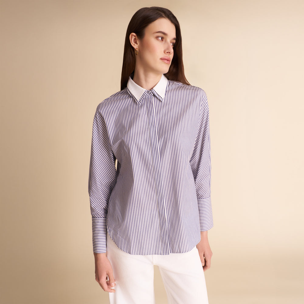 A woman modeling a long-sleeve button-up shirt with thin vertical blue and white stripes, featuring a solid white collar, paired with white trousers against a solid beige studio background.