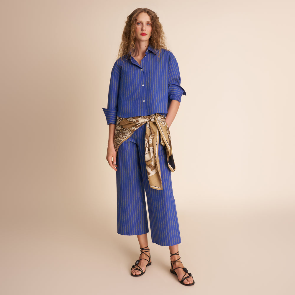 Woman wearing a blue striped shirt and wide-leg pants with a gold scarf, standing against a beige background.