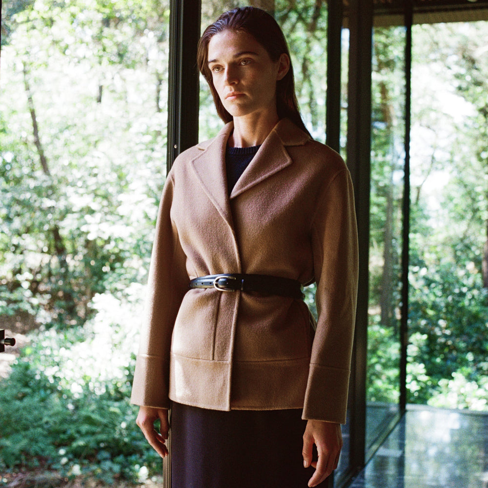 Woman wearing a beige coat standing in front of a glass door with a forest view.