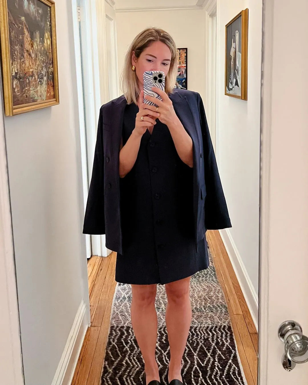 Woman taking a mirror selfie wearing a black blazer and dress in a hallway.