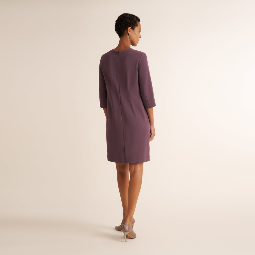 A woman stands with her back to the camera wearing a knee-length mauve dress with elbow-length sleeves and nude high heels, photographed against a plain warm off-white studio background.