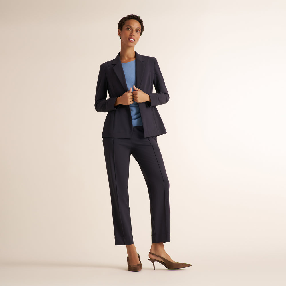 A woman modeling a dark navy blue tailored pantsuit, consisting of a single-breasted blazer and matching trousers, styled with a light blue top and brown slingback heels against a solid cream studio background.