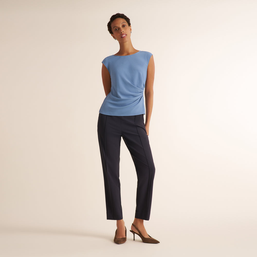 A model stands against a warm off-white studio background wearing a light blue sleeveless top with a draped side detail and dark navy cropped trousers, paired with brown pointed-toe heels.