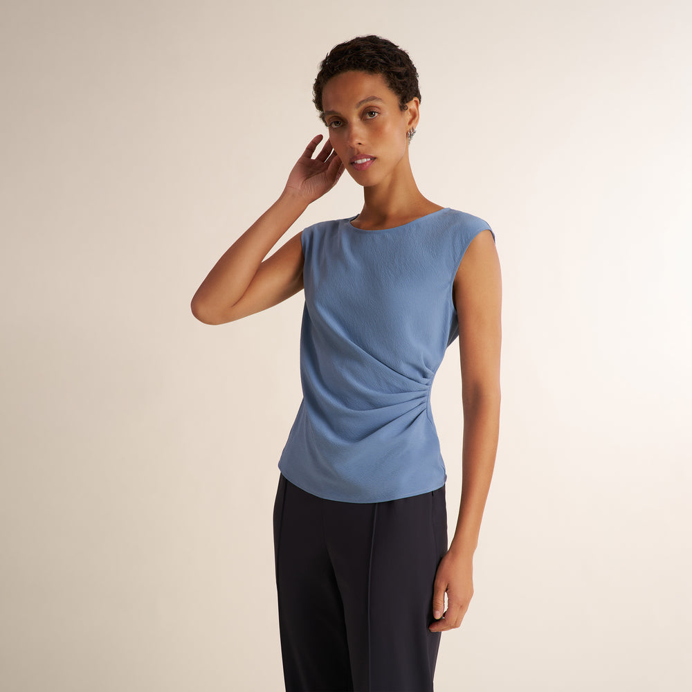 A woman modeling a light blue sleeveless knit top with a draped side detail, paired with dark trousers, against a plain beige studio background.