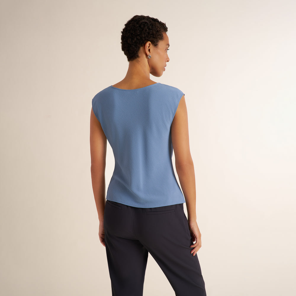 A woman shown from the back wearing a light blue sleeveless top and dark navy pants, standing against a plain light beige studio background.