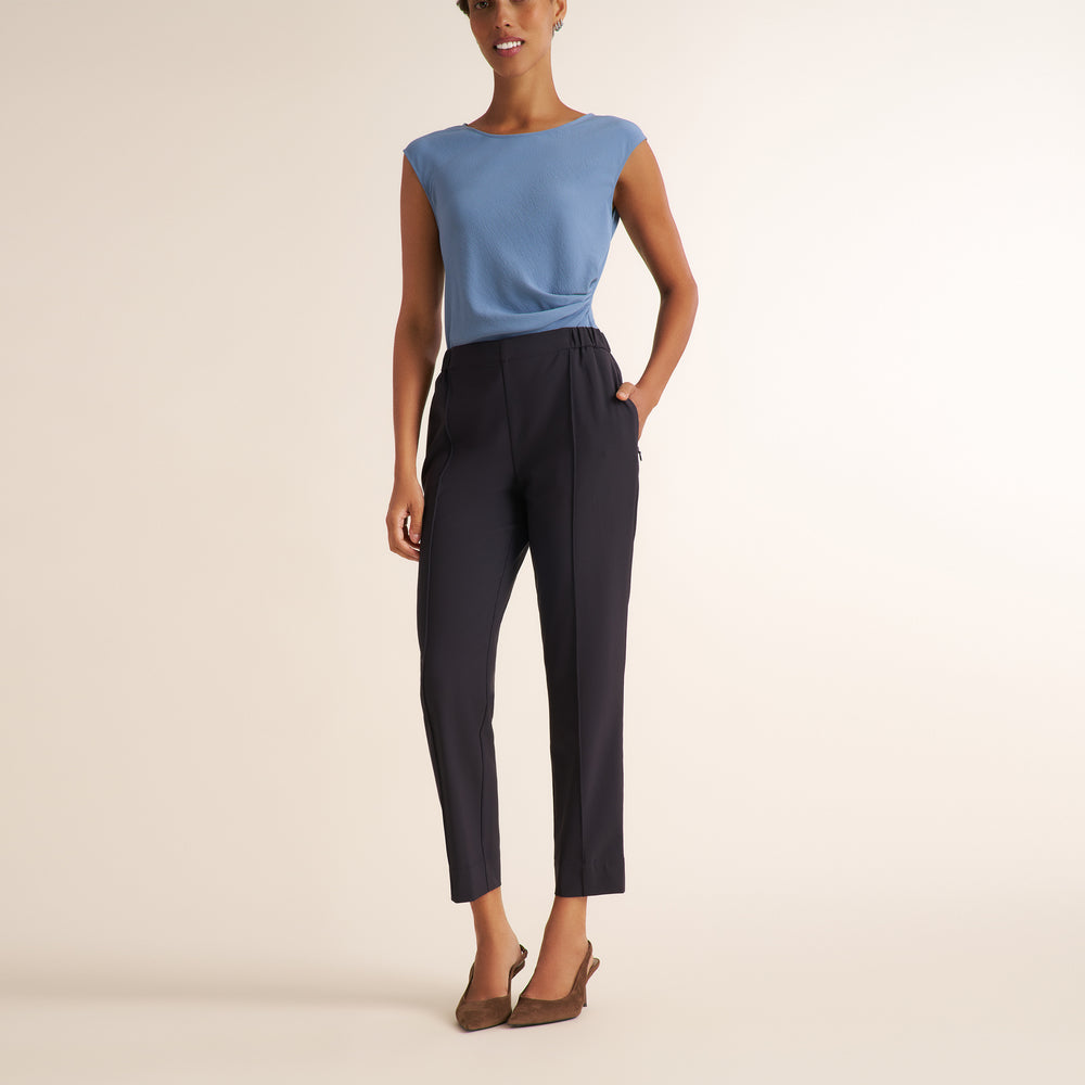A model wearing a light blue sleeveless top and dark charcoal cropped trousers, paired with brown pointed-toe flats, against a solid warm beige studio background.