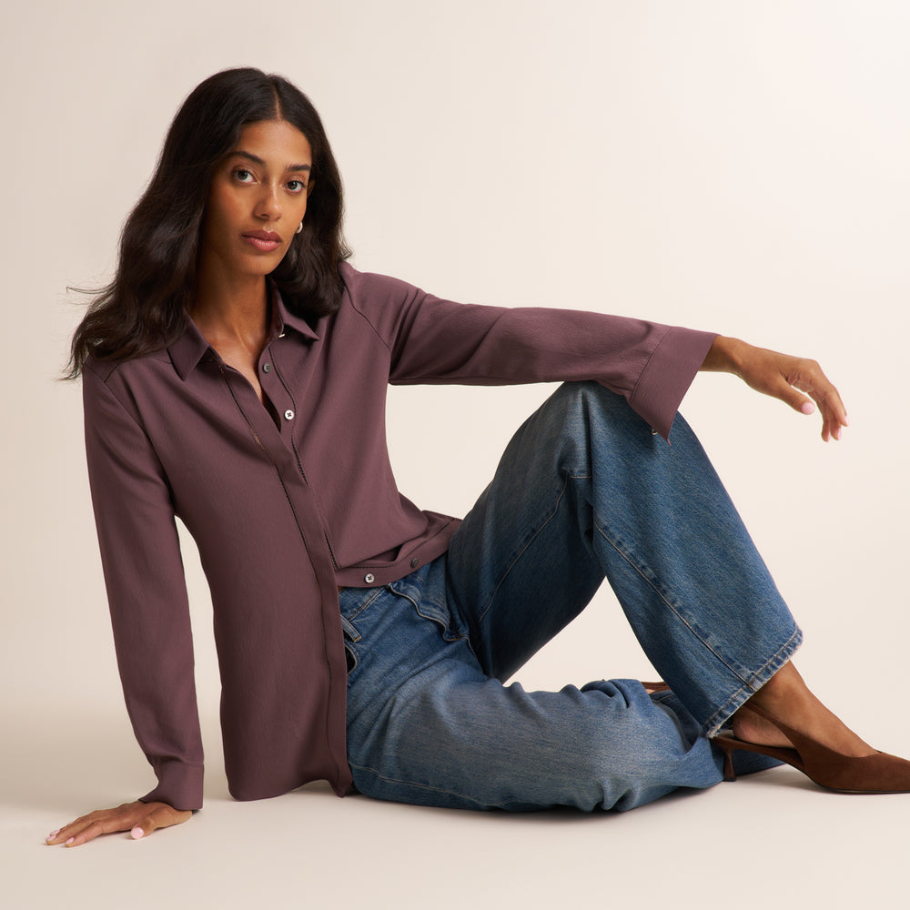A woman wearing a long-sleeve button-up shirt in a muted plum purple and blue jeans, sitting on a plain off-white studio background.