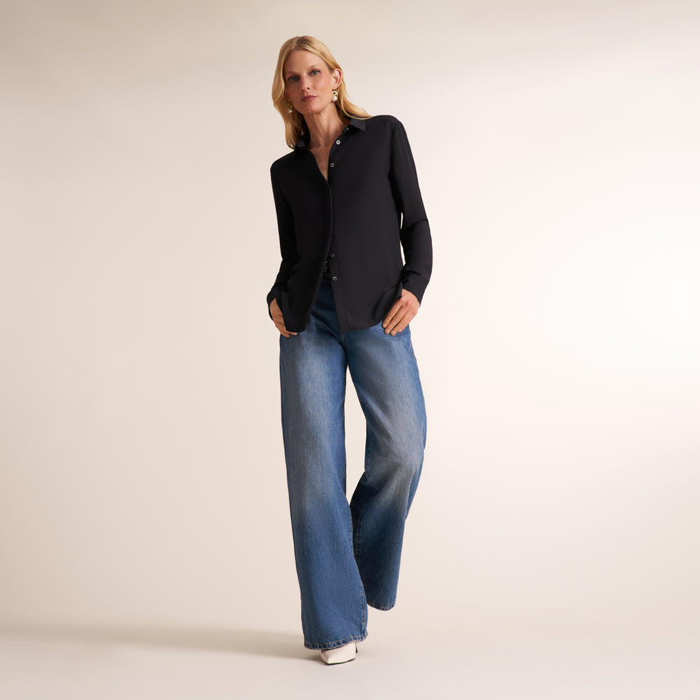 A woman modeling a black long-sleeve button-up shirt and wide-leg blue jeans against a plain light beige studio background.