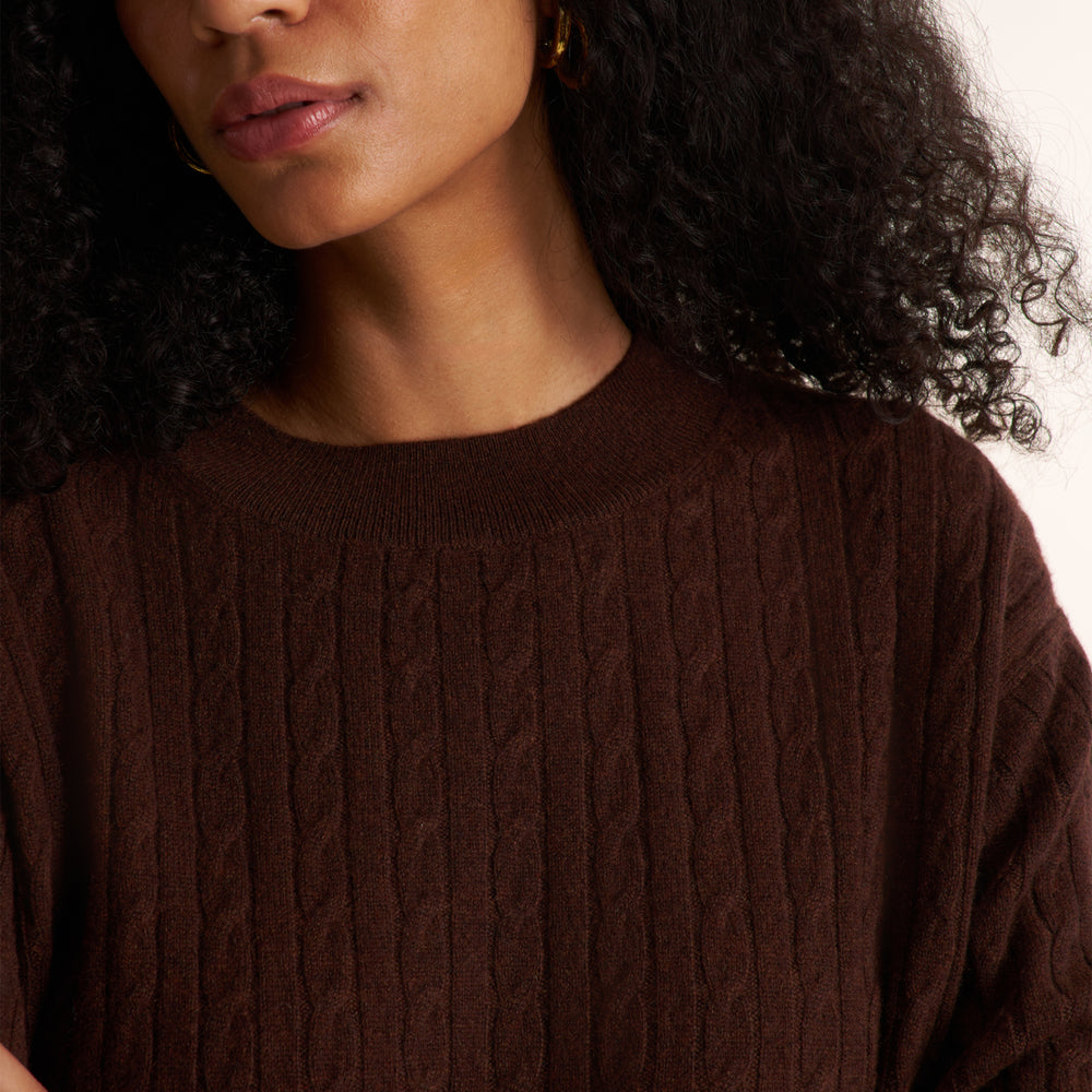 A close-up shot of a woman wearing a dark brown cable-knit crewneck sweater, featuring her curly hair and a neutral background.