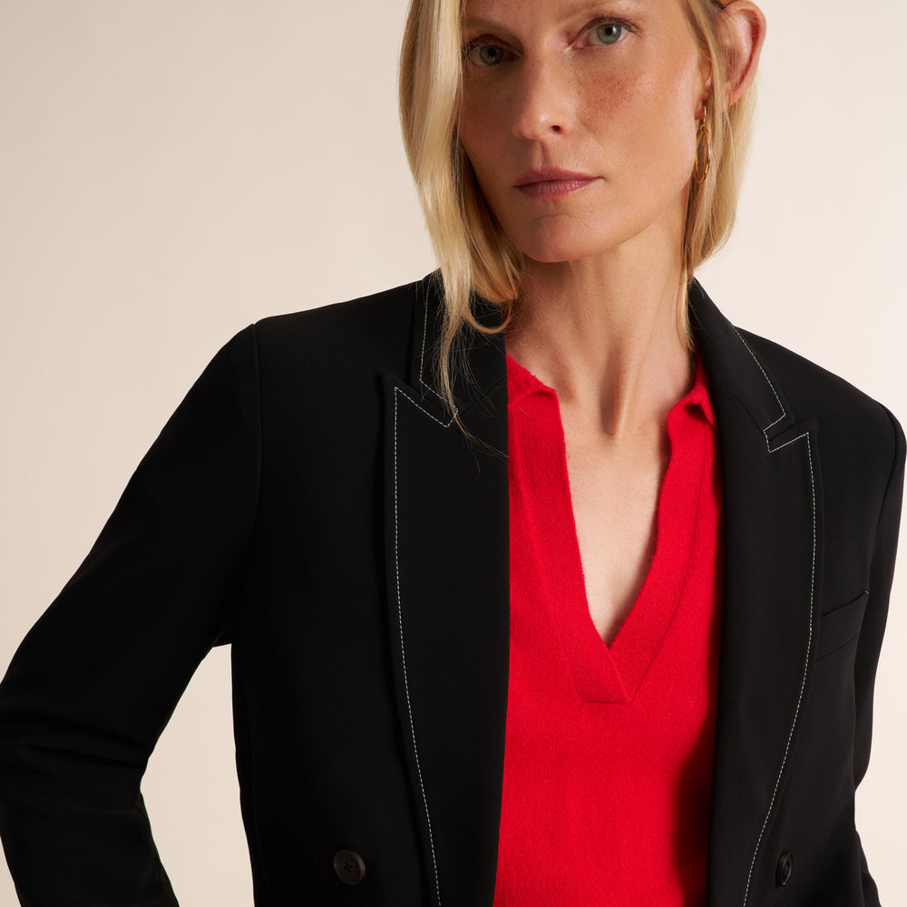 A close-up of a woman wearing a black blazer with white contrast stitching over a bright red V-neck top, set against a plain beige background.