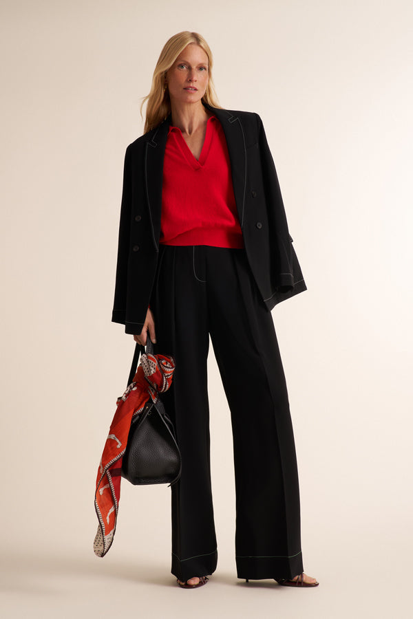 A woman models a black double-breasted blazer with wide-leg black trousers, a vibrant red V-neck top, and holds a black handbag with a red patterned scarf, set against a solid light beige background.