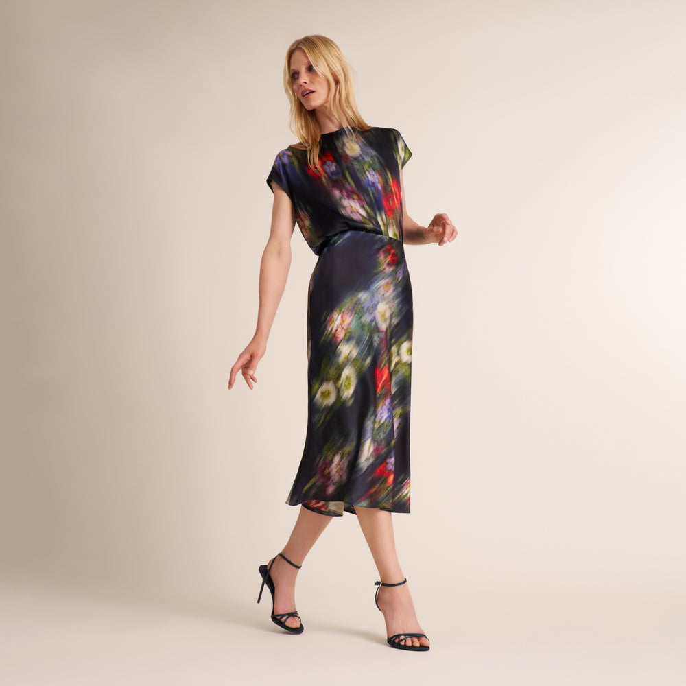 A woman modeling a short-sleeve midi dress with a dark base and a colorful abstract floral print, paired with black high-heeled sandals, against a plain beige studio background.