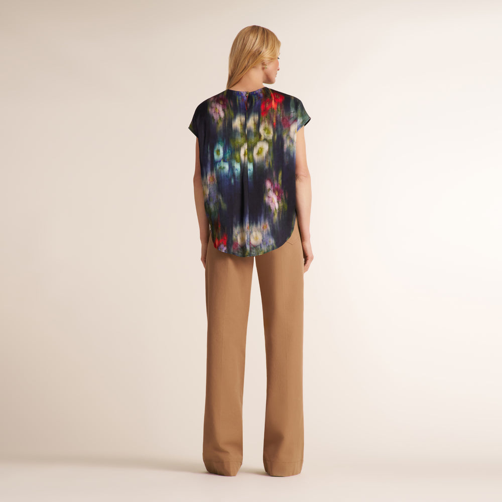 A woman stands with her back to the camera wearing a short-sleeve top with a dark abstract floral print and tan wide-leg pants against a plain beige studio background.