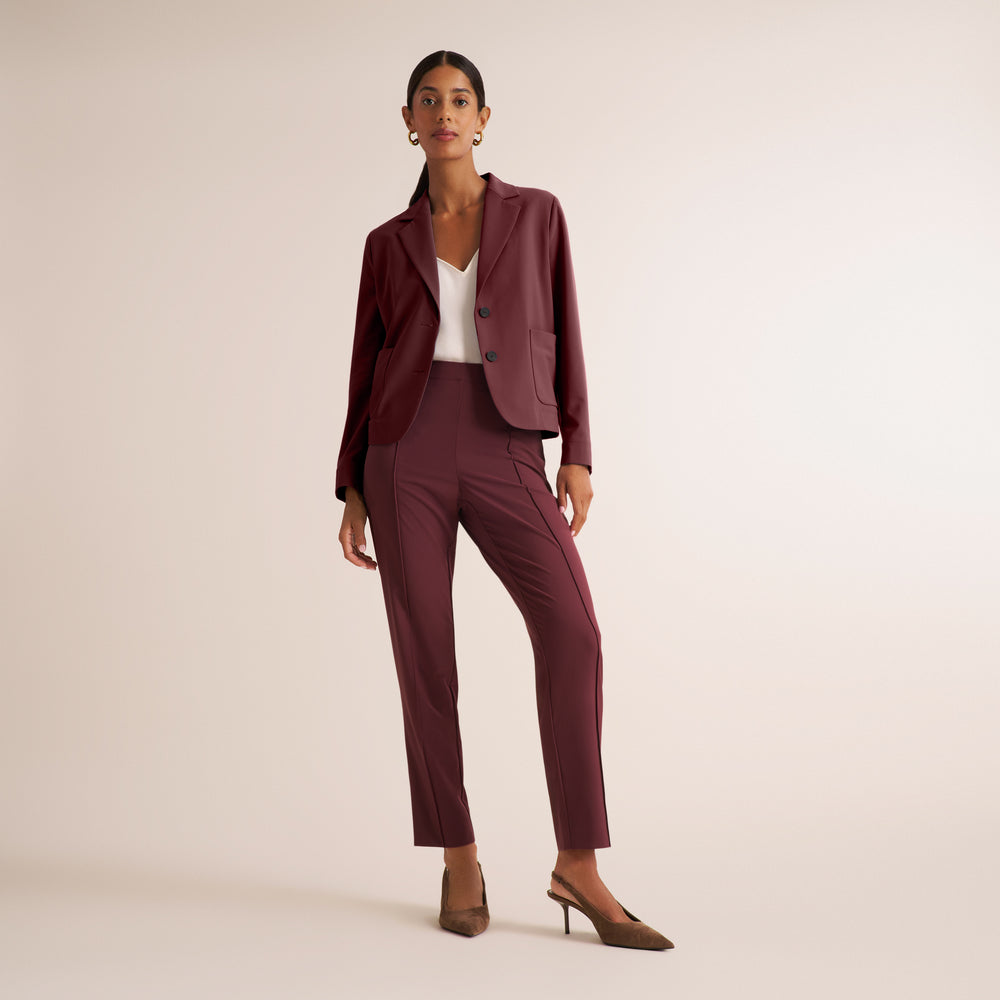 A model stands in a studio wearing a matching burgundy blazer and trousers set with a white top and brown pointed-toe heels, against a plain light beige background.
