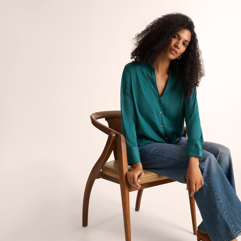 A woman with curly black hair sits on a wooden chair wearing a teal green satin button-up blouse and blue jeans against a warm off-white studio background.