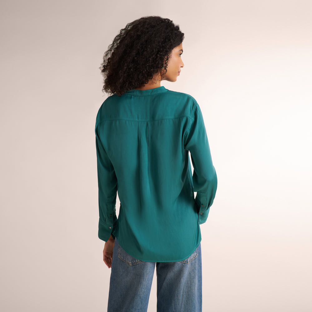 A woman with curly dark hair is shown from the back wearing a teal green long-sleeve blouse and blue jeans against a plain light beige studio background.