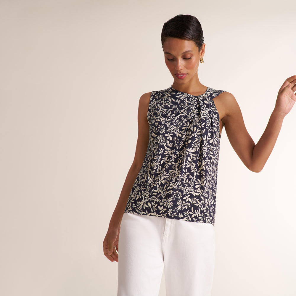 A woman modeling a sleeveless top with a navy blue and white floral pattern, paired with white pants, against a plain beige studio background.
