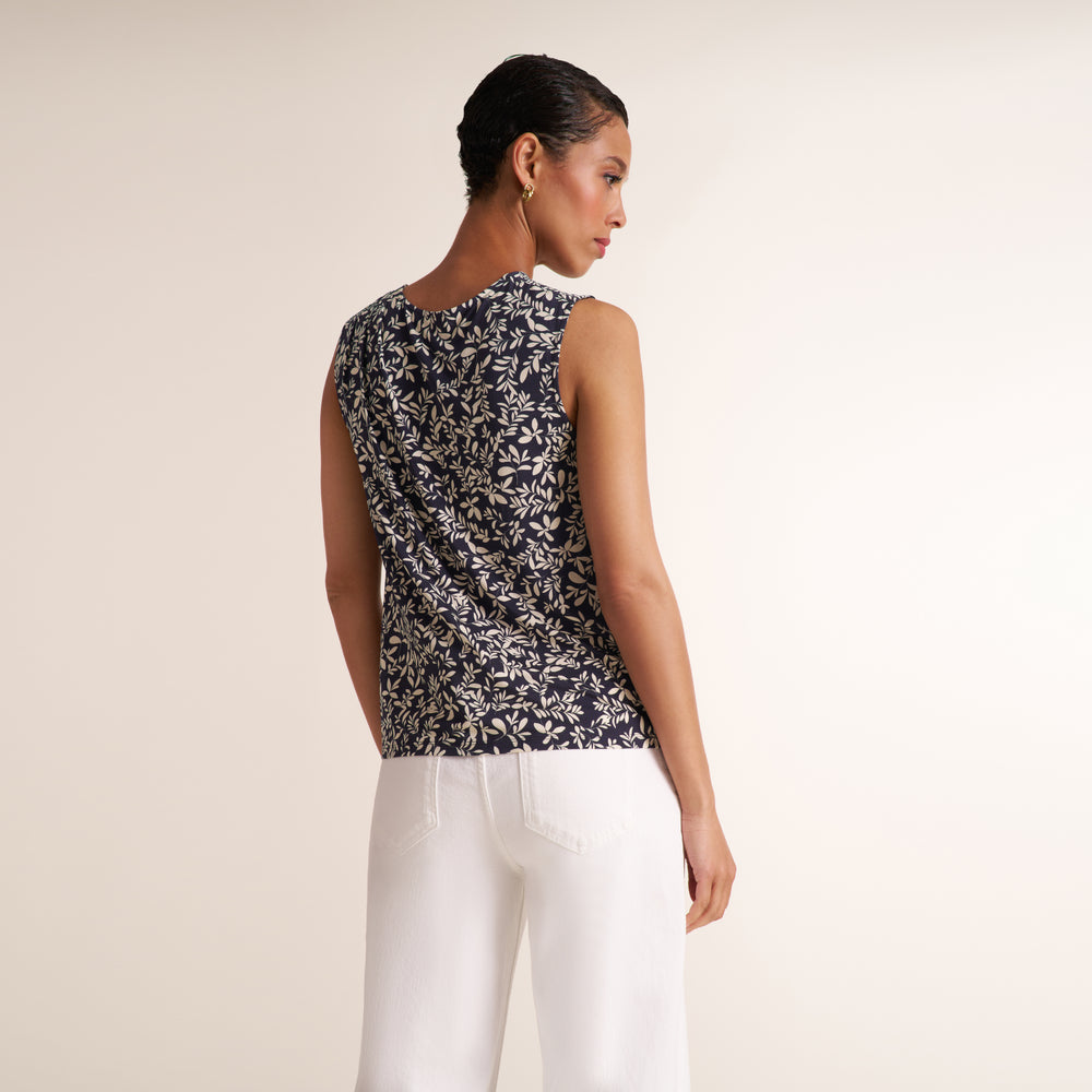 A woman shown from the back wearing a sleeveless black and white floral print top and white pants, standing against a plain light beige studio background.