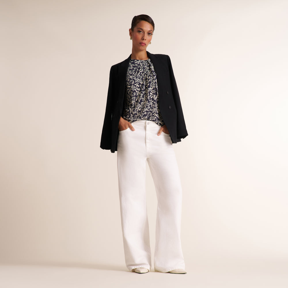 A model stands against a plain beige studio background wearing a black blazer, a black-and-white patterned top, and white wide-leg trousers.