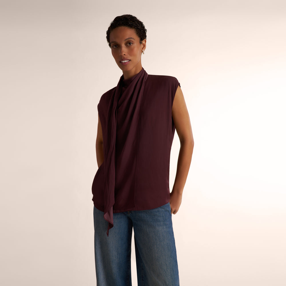 A model shown wearing a sleeveless, high-neck, dark burgundy top and blue jeans, standing against a plain light beige studio background.