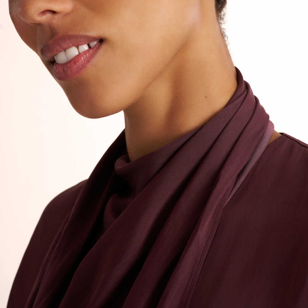 A model shown wearing a sleeveless, high-neck, dark burgundy top and blue jeans, standing against a plain light beige studio background.