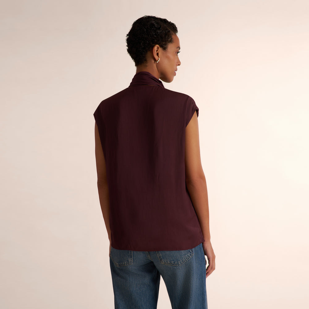 A model shown from the back wearing a sleeveless, high-neck, dark burgundy top and blue jeans, standing against a plain light beige studio background.