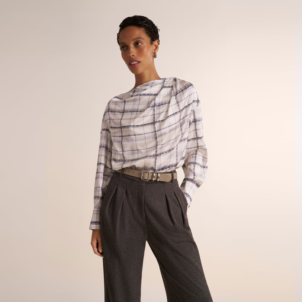 A woman modeling a white and gray plaid shirt, paired with a gray herringbone blazer and matching pants, against a plain light beige studio background.