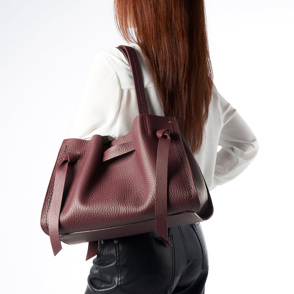 A woman wearing a white blouse and black pants carries a large pebbled leather shoulder bag in a deep burgundy shade against a plain white background.