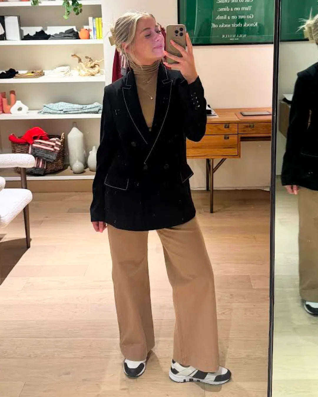 Person taking a mirror selfie wearing a black blazer and beige pants in a store.