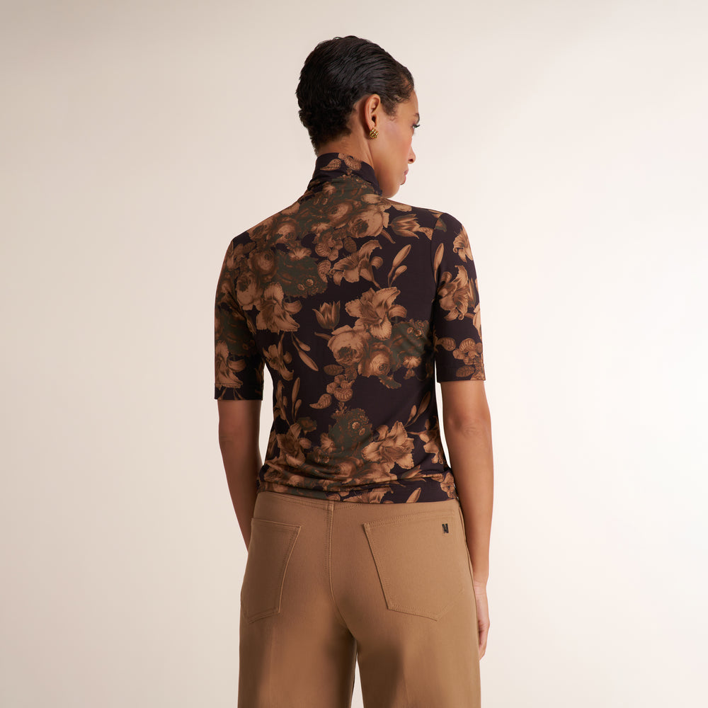 A woman shown from the back wearing a fitted short-sleeve mock-neck top with a dark floral print in brown and olive tones, paired with tan pants, against a plain light beige studio background.