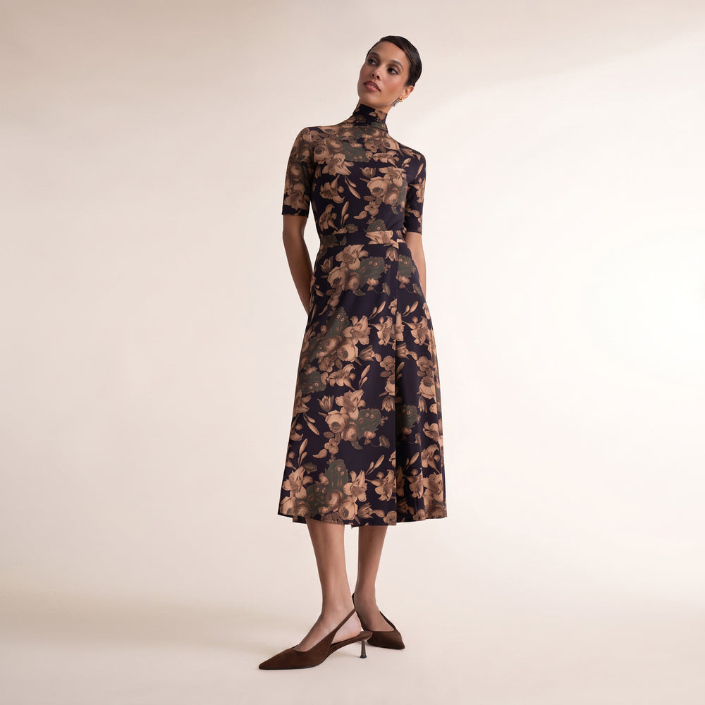 A woman modeling a short-sleeved midi dress with a high mock neck and an all-over floral print in beige and brown tones on a dark navy base, paired with dark brown pointed-toe slingback heels, against a plain cream studio background.