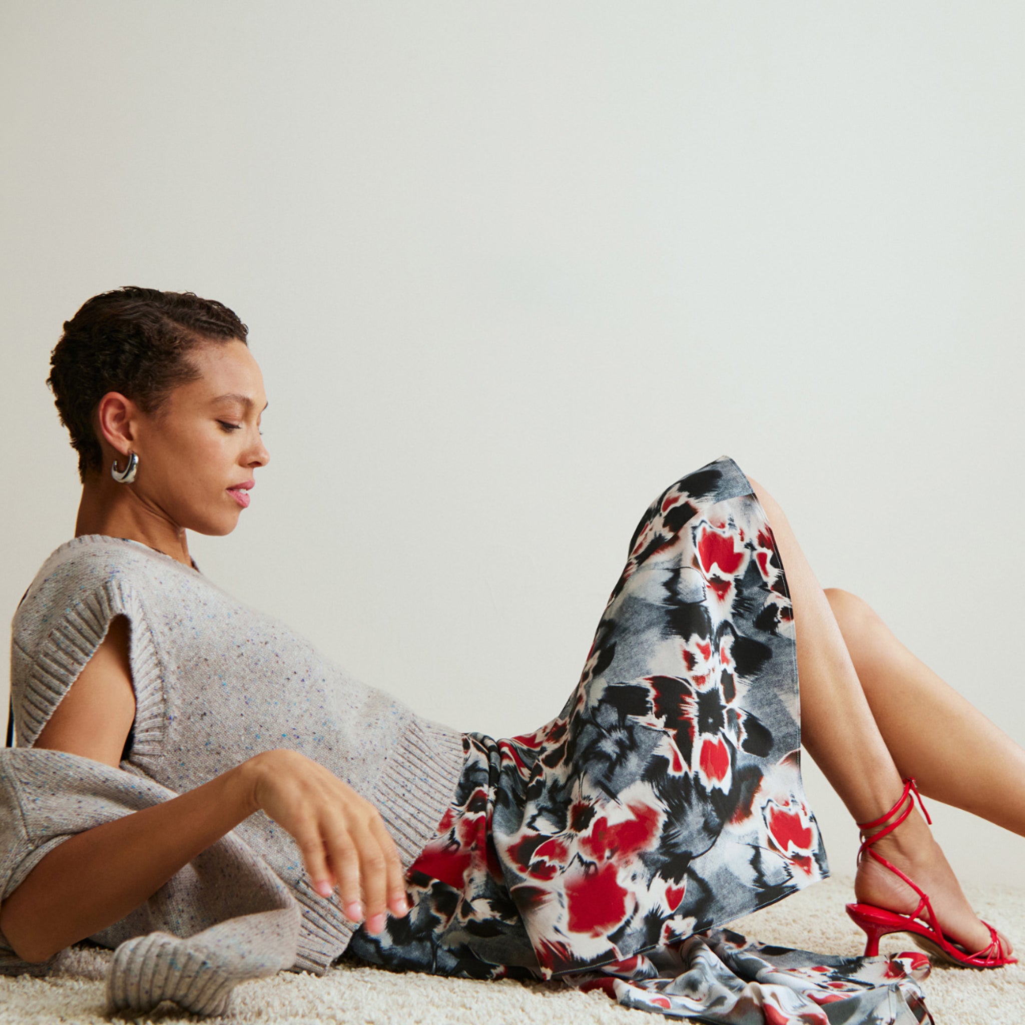 Woman sitting on a carpet with a floral-patterned blanket draped over her legs, wearing a gray sweater and red sandals.