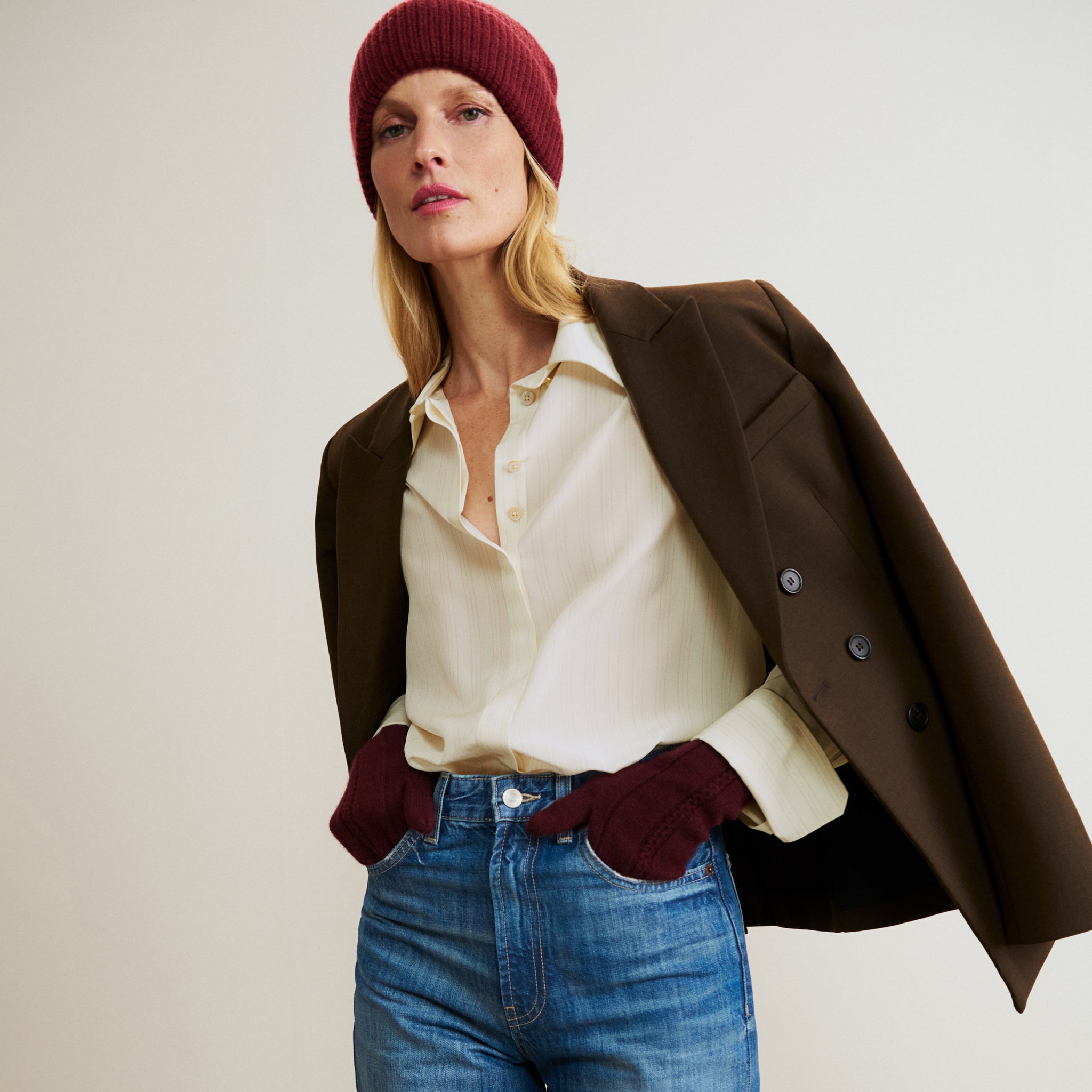 Woman wearing a brown coat, white shirt, blue jeans, and red beanie and gloves on a plain background