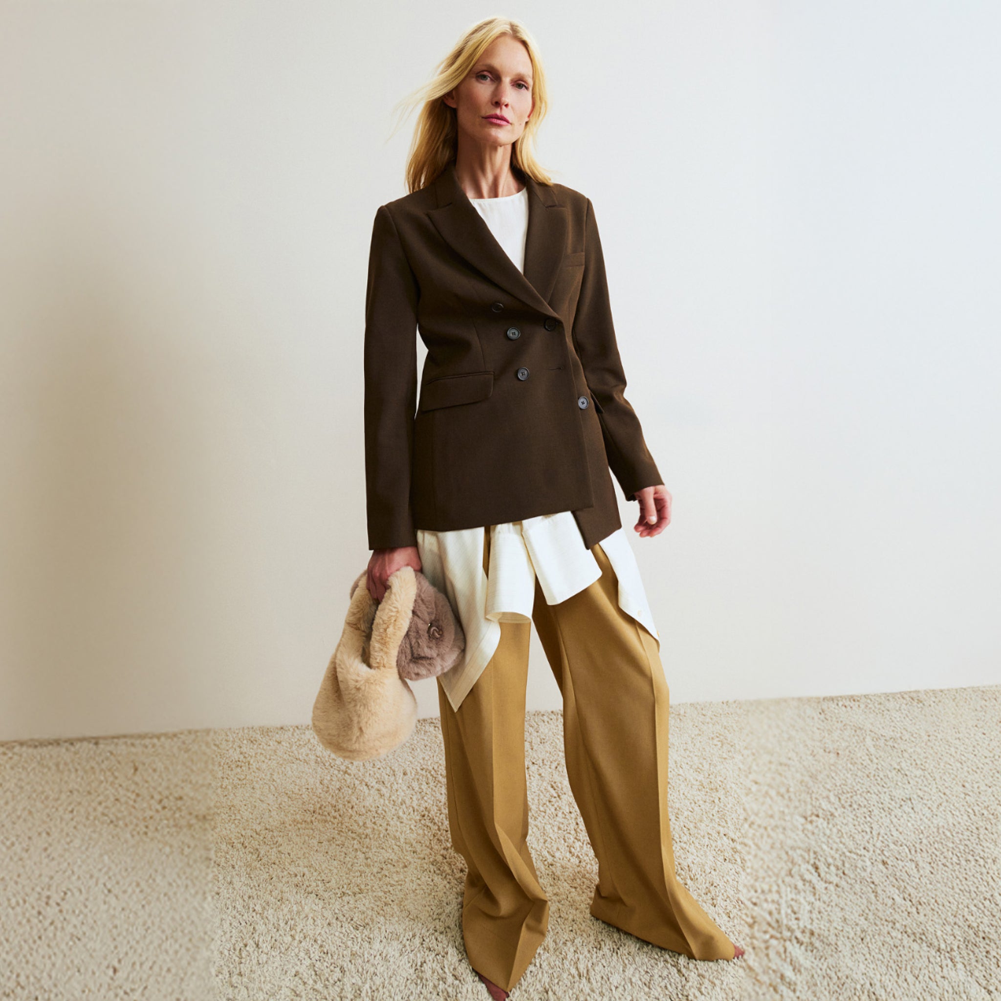 Woman wearing a brown blazer and beige pants holding a beige handbag on a plain background