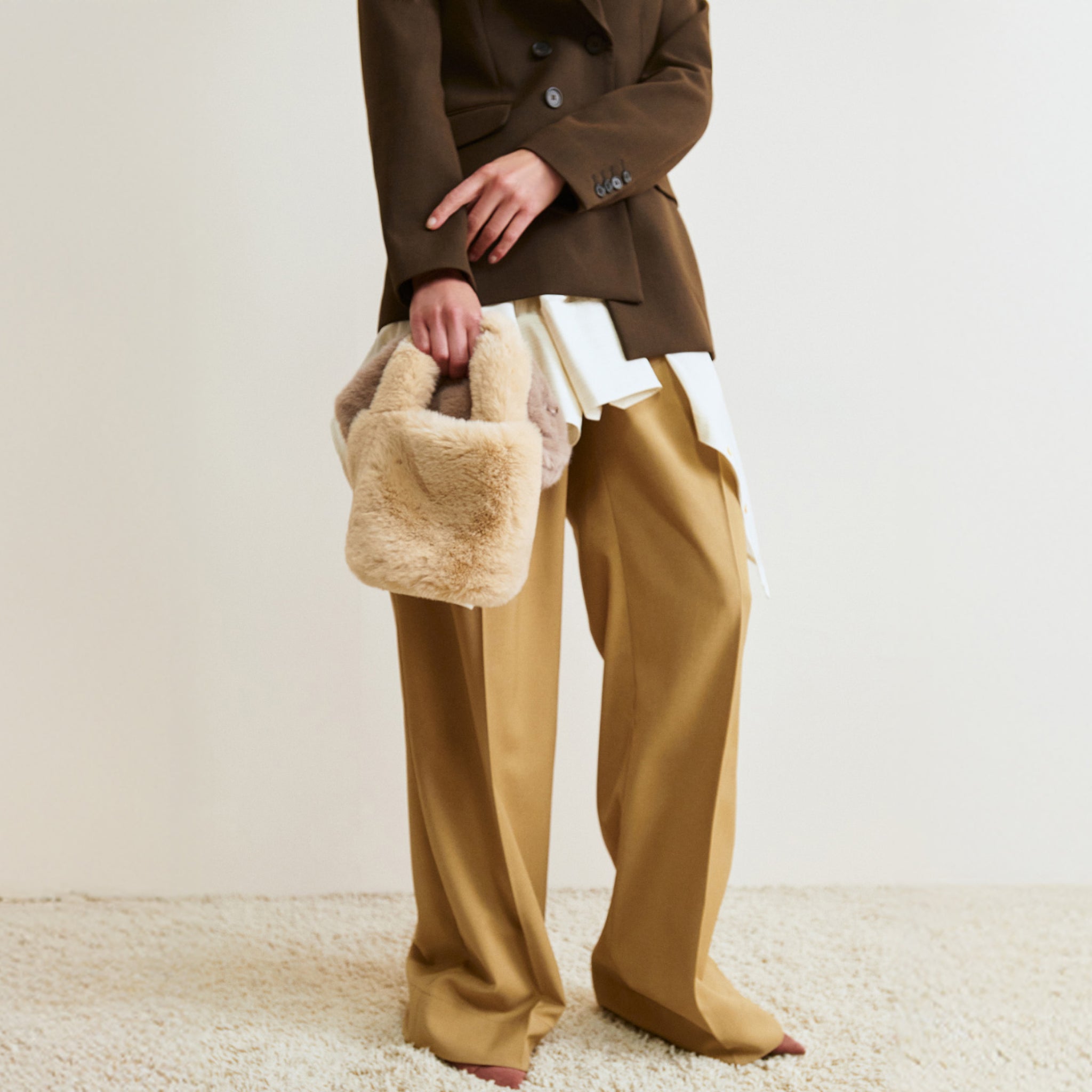 Person wearing a brown coat and gold pants holding a beige bag on a neutral background