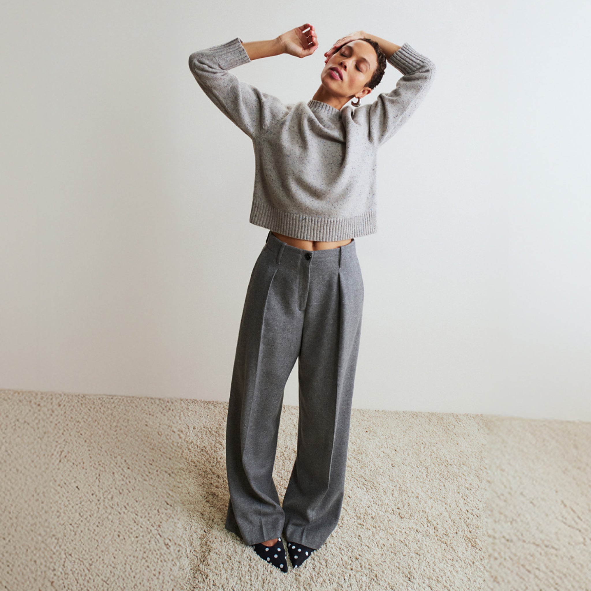 Person wearing a gray sweater and pants standing on a beige carpet with a white wall background