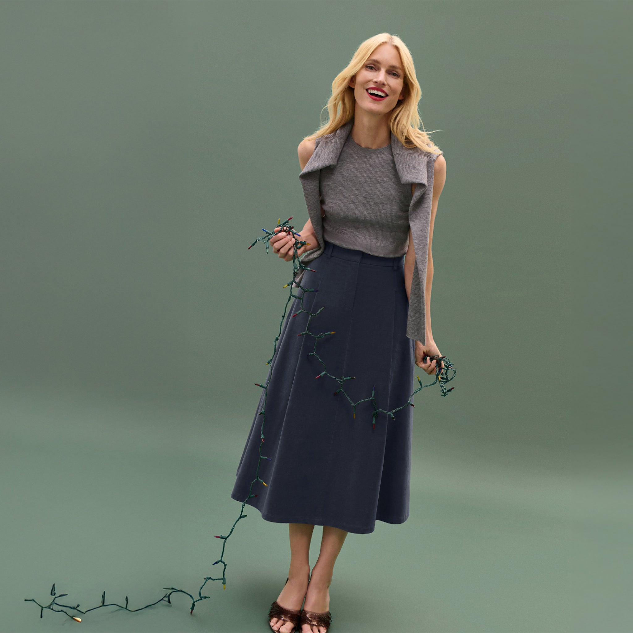 Woman wearing a gray vest and navy skirt holding greenery against a green background