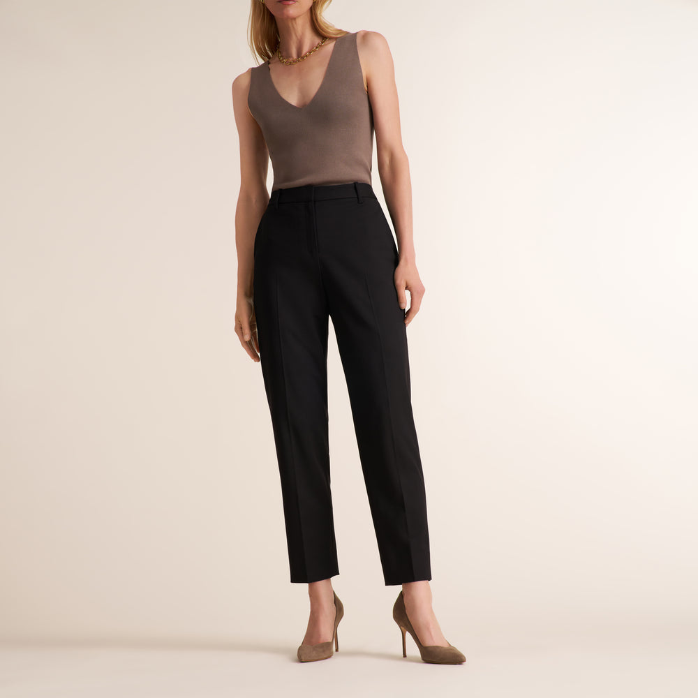 A model wearing black cropped trousers with a V-neck taupe knit top and beige heels, standing against a plain beige studio background.