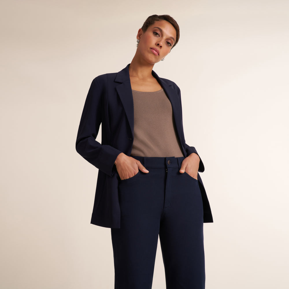 A woman modeling a navy blue tailored blazer and matching trousers over a taupe top, photographed against a plain light beige studio background.