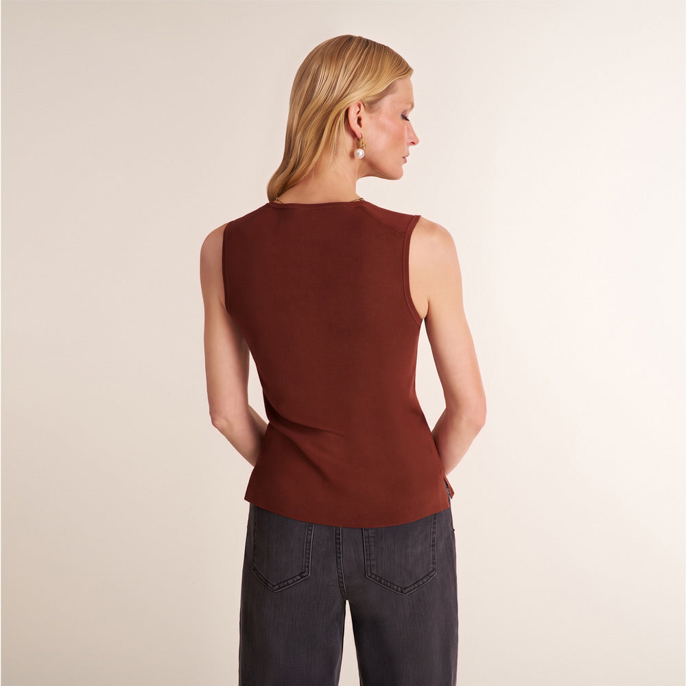 A woman shown from the back wearing a sleeveless rust-brown top and dark gray jeans, standing against a plain light beige studio background.