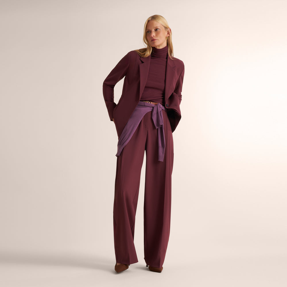 Woman wearing an aubgerine ribbed turtleneck with matching wide leg pants and blazer with a fig purple sweater around her waist