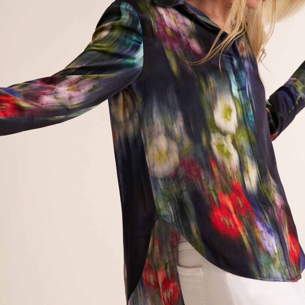 A woman wearing a long-sleeve button-up blouse with a dark navy base and a colorful painterly floral print, paired with white pants against a plain light background.