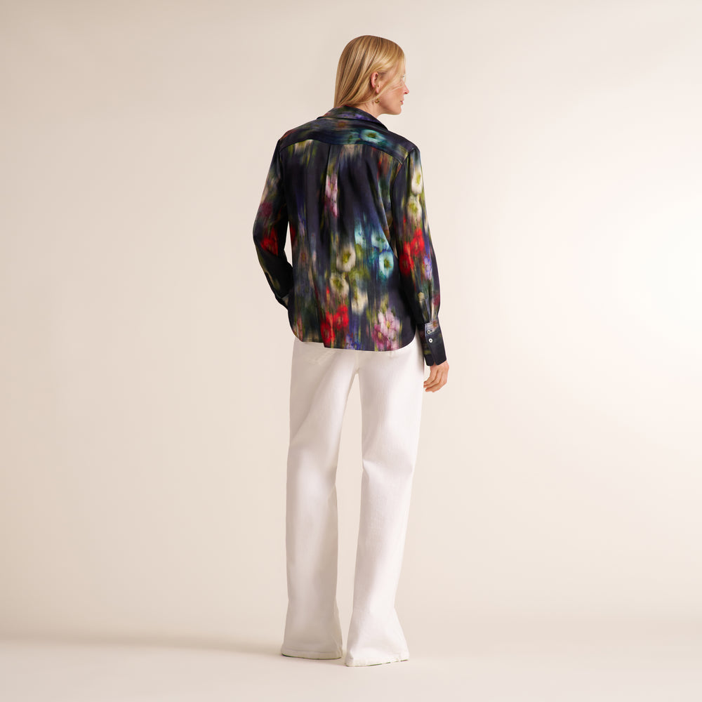 A woman stands with her back to the camera wearing a long-sleeve button-up shirt with a dark abstract multicolor print and white wide-leg trousers, set against a plain light beige studio background.